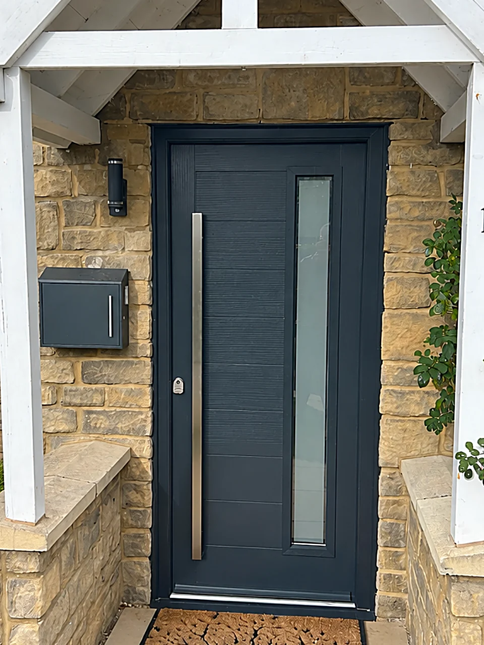 Contemporary anthracite grey composite front door with a full-height obscure glazed panel and long stainless steel bar handle, beneath a white timber porch on a Cotswold stone house, Cricklade