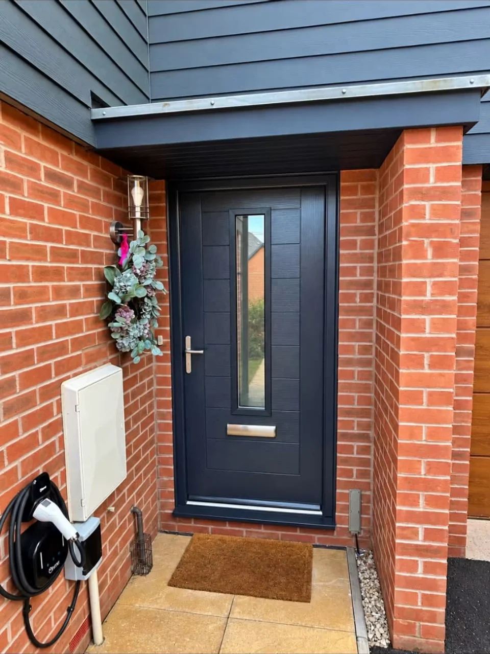 Dark composite front door with vertical glazed panel installed in a brick porch in Innsworth, Gloucestershire